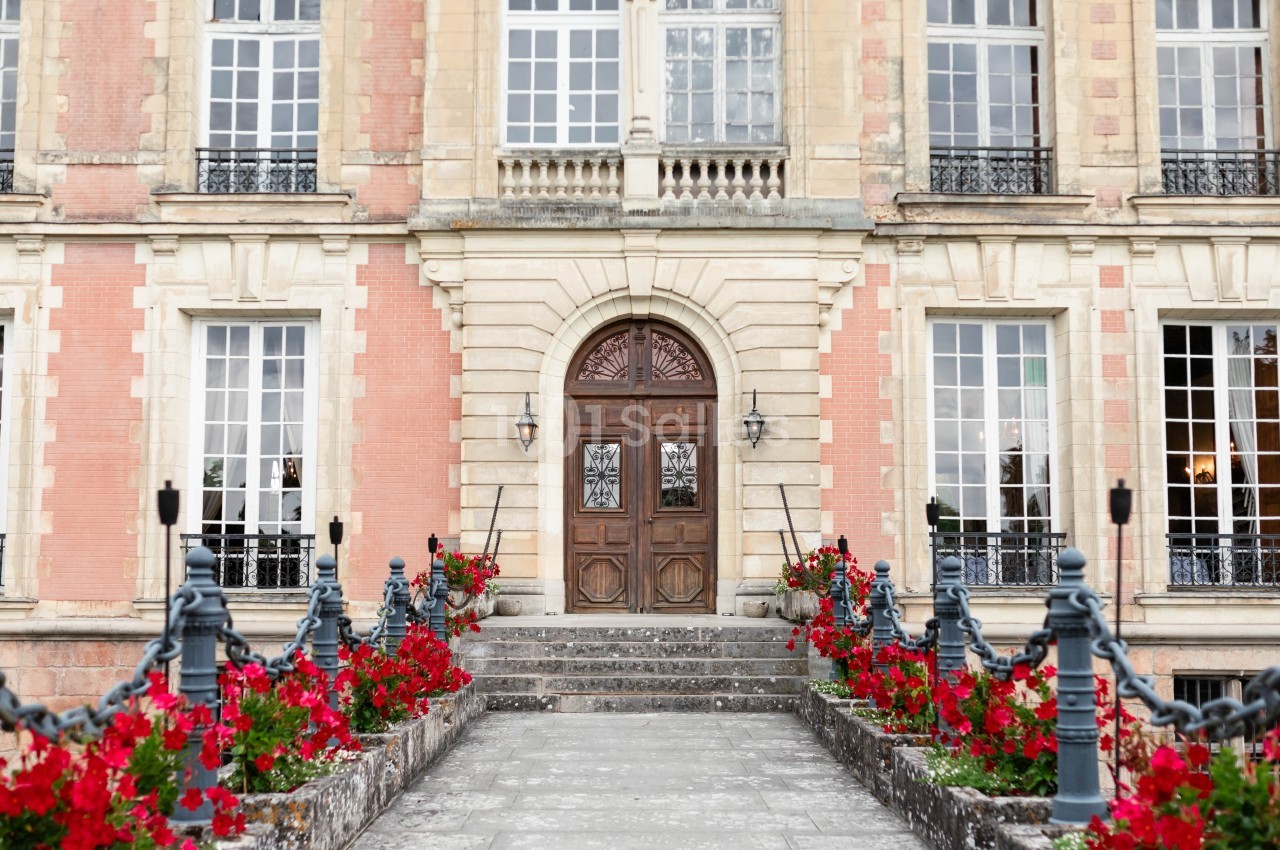 Façade d'un bâtiment ancien avec porte en bois sculpté, grandes fenêtres et allée bordée de fleurs rouges.