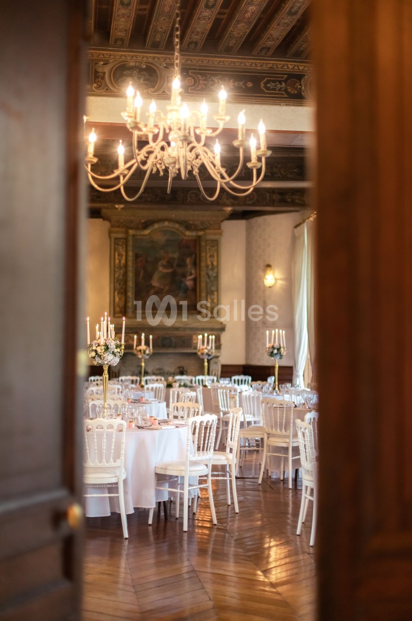 Salle de réception élégante avec tables dressées, chaises blanches et lustre suspendu sous un plafond décoré.