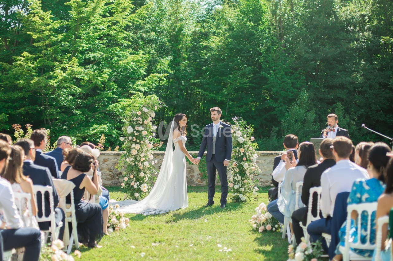 Un couple échange ses vœux lors d'une cérémonie de mariage en plein air, entouré d'invités et de décorations florales.