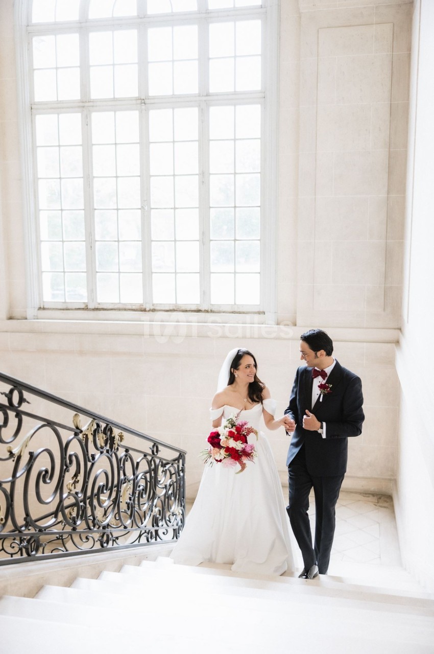 Un couple de mariés descend un escalier élégant dans un bâtiment lumineux avec une grande fenêtre en arrière-plan.