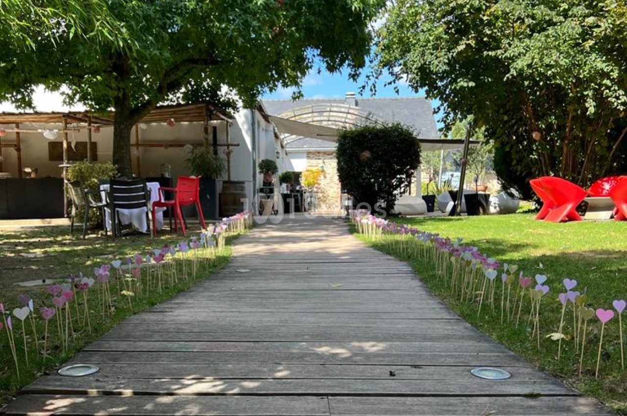 Allée en bois bordée de petits cœurs colorés plantés dans l'herbe, menant à une terrasse ombragée.