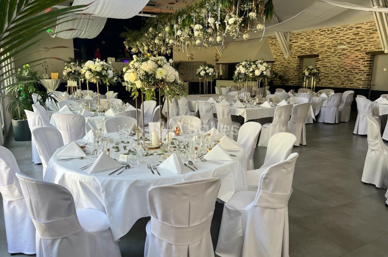 Salle de réception décorée avec des tables rondes, nappes blanches, chaises houssées et arrangements floraux suspendus.