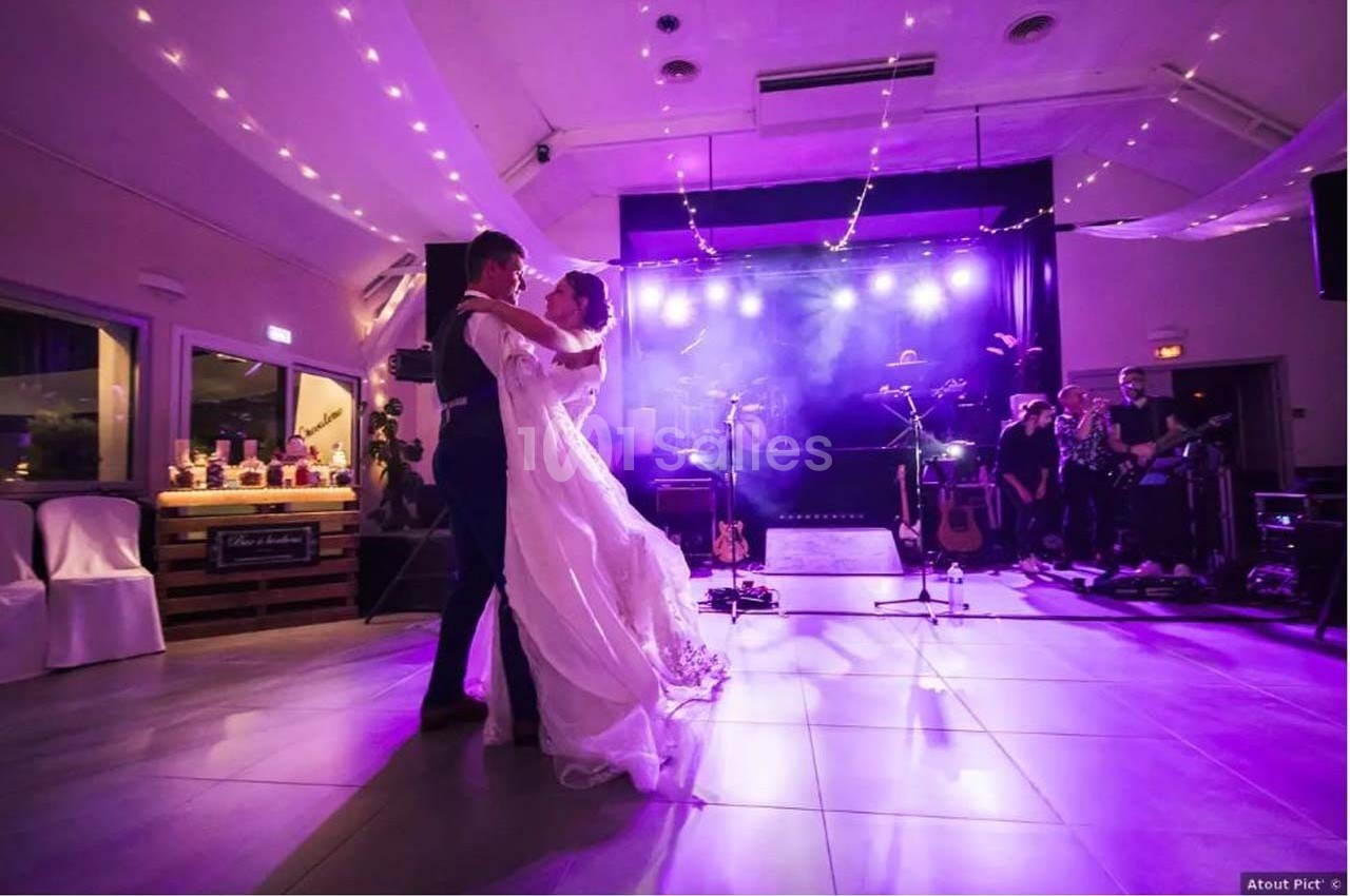 Un couple danse sous des lumières violettes dans une salle décorée, avec des musiciens en arrière-plan.