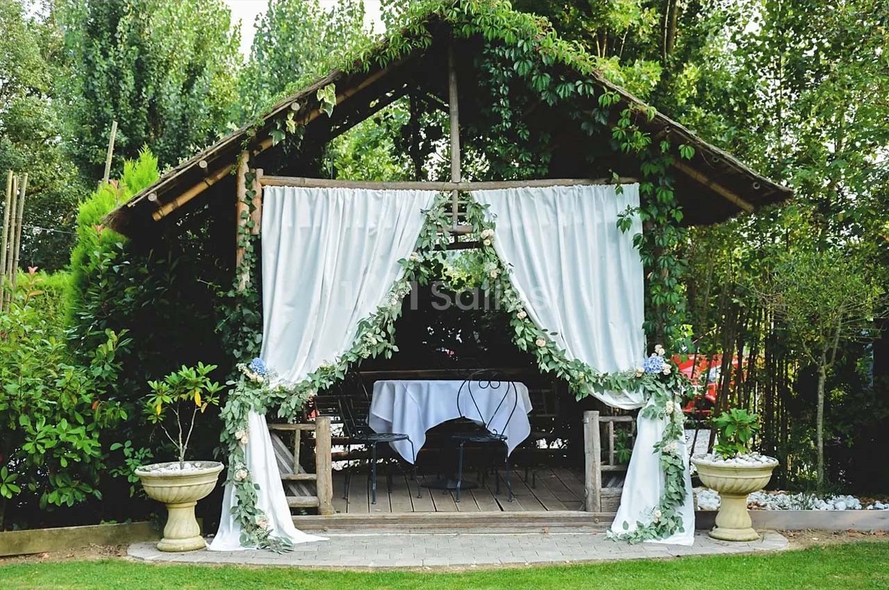 Abri en bois décoré de rideaux blancs et de feuillage, abritant une table dressée dans un jardin verdoyant.
