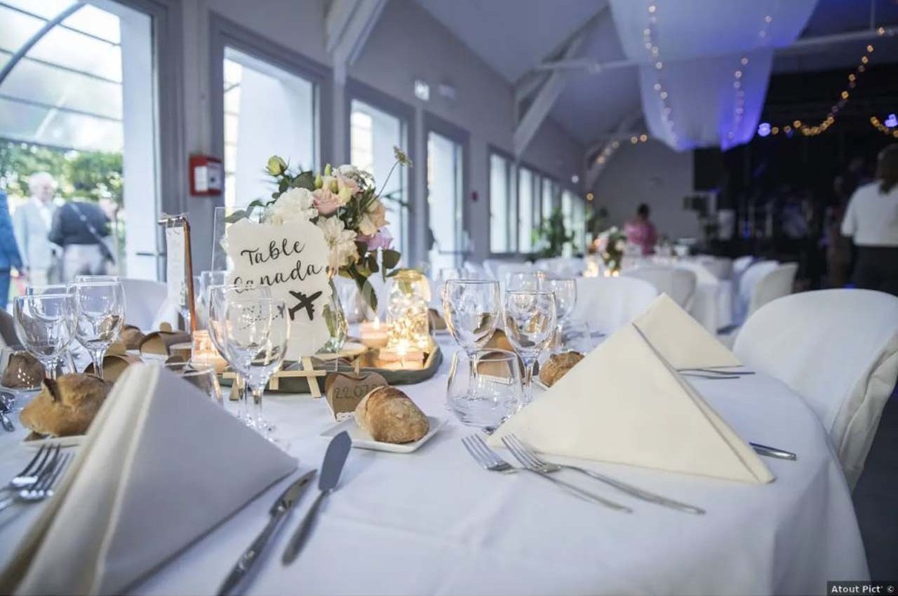Table décorée pour un mariage avec nappes blanches, serviettes pliées, fleurs, bougies et marque-place ’Table Canada’.