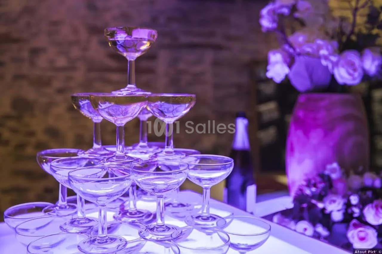 Pyramide de coupes à champagne éclairée en violet, avec un vase de fleurs et une bouteille en arrière-plan.