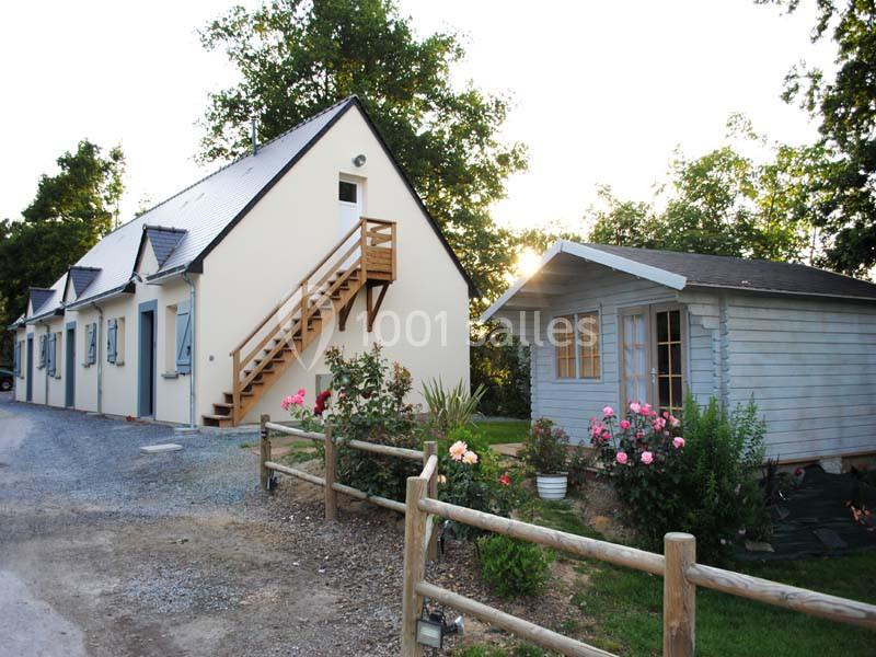 Maison blanche à étage avec escalier extérieur en bois, cabanon en bois à côté, jardin fleuri et clôture en bois.