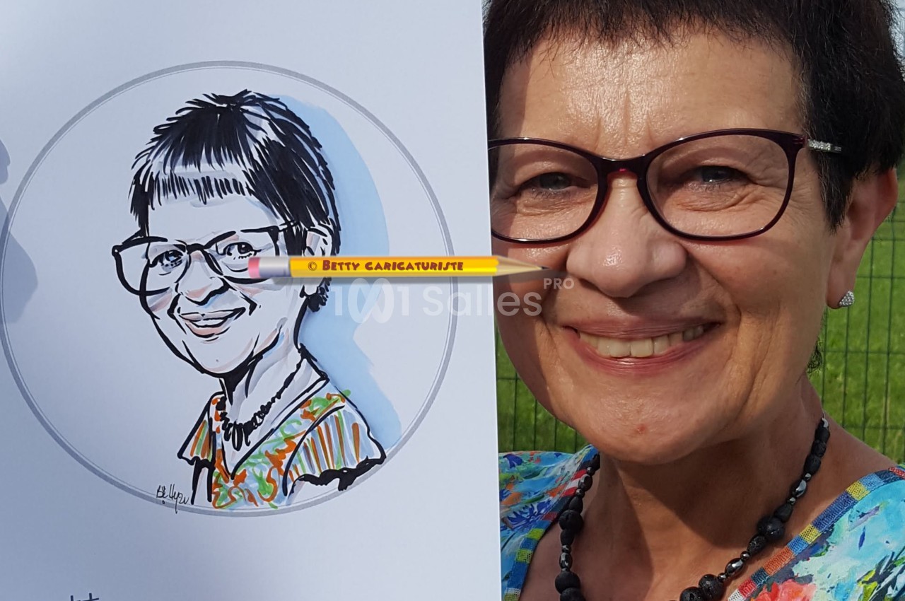 Femme souriante tenant un dessin caricatural d'elle-même, en extérieur devant une clôture et un ciel dégagé.