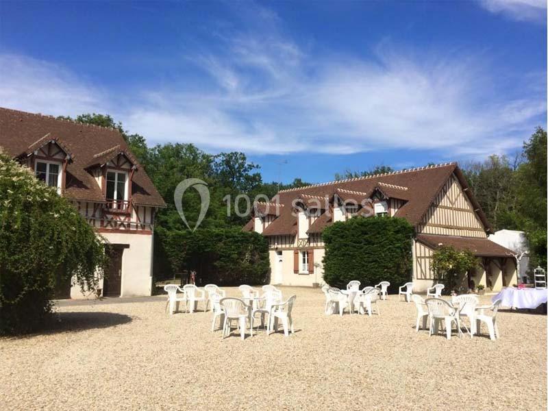 Cour d'une maison à colombages avec tables et chaises blanches en plastique, entourée de verdure sous un ciel bleu.
