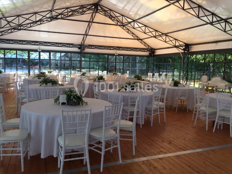 Salle couverte avec des tables rondes dressées, nappes blanches et chaises, entourée de baies vitrées et de verdure.
