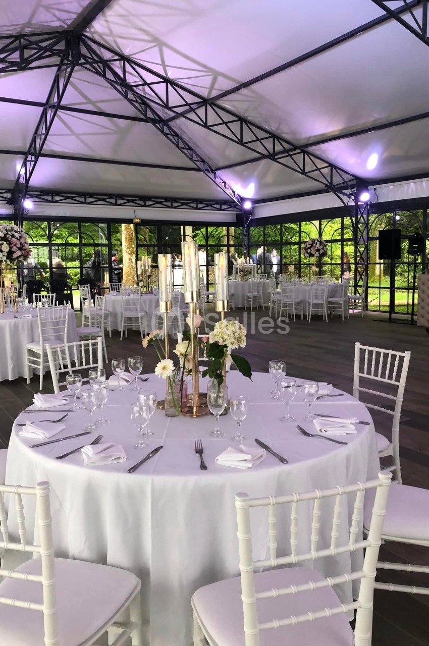 Salle de réception sous une structure en toile avec tables rondes dressées, décor floral et lumière naturelle.