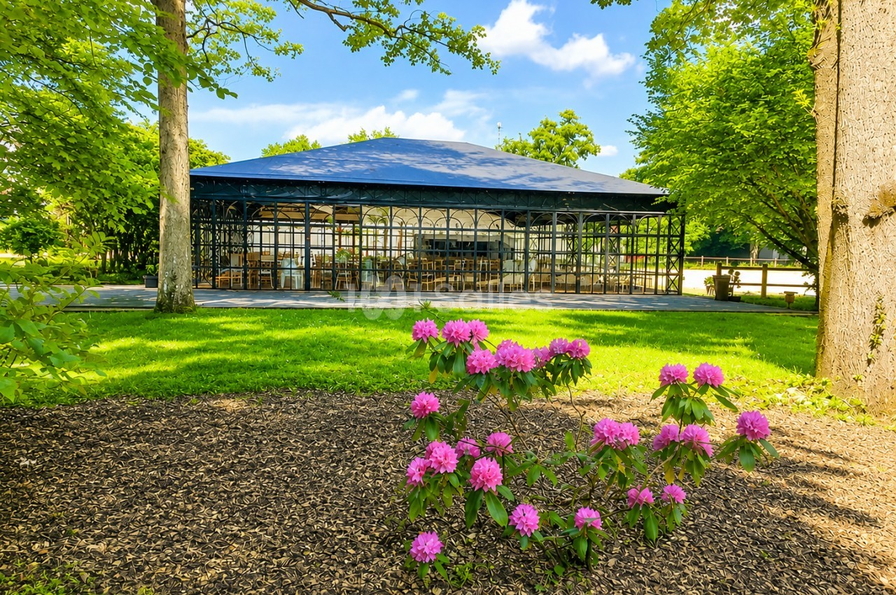 Serre vitrée entourée de verdure avec des fleurs de rhododendrons roses au premier plan.