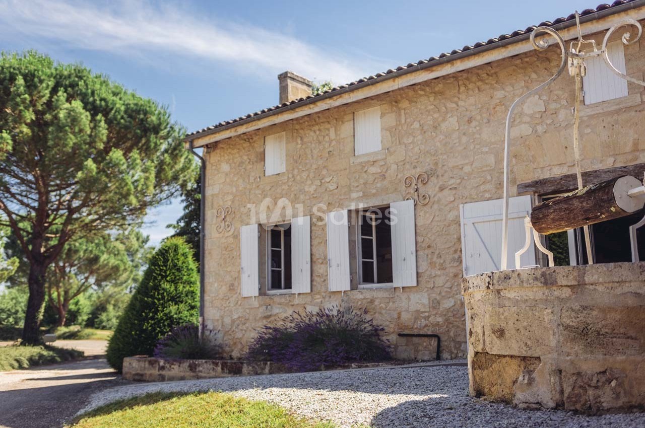 Maison en pierre avec volets blancs, entourée de végétation, avec un puits en pierre au premier plan.