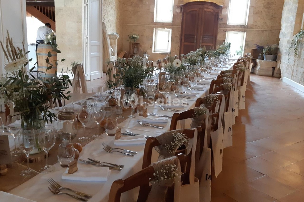 Grande table de banquet dressée avec nappes blanches, décorations florales et sacs cadeaux dans une salle en pierre.
