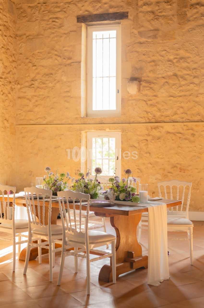 Table en bois décorée de fleurs et entourée de chaises blanches dans une salle en pierre avec lumière naturelle.
