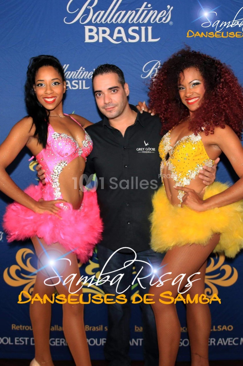 Un homme pose avec deux danseuses en costumes colorés devant un fond promotionnel ’Ballantine's Brasil’.
