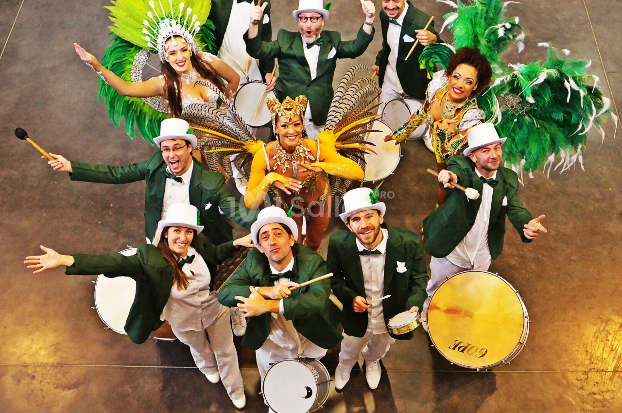 Groupe de musiciens en costumes verts et blancs accompagnés de danseuses en tenues colorées et plumes, vu de dessus.