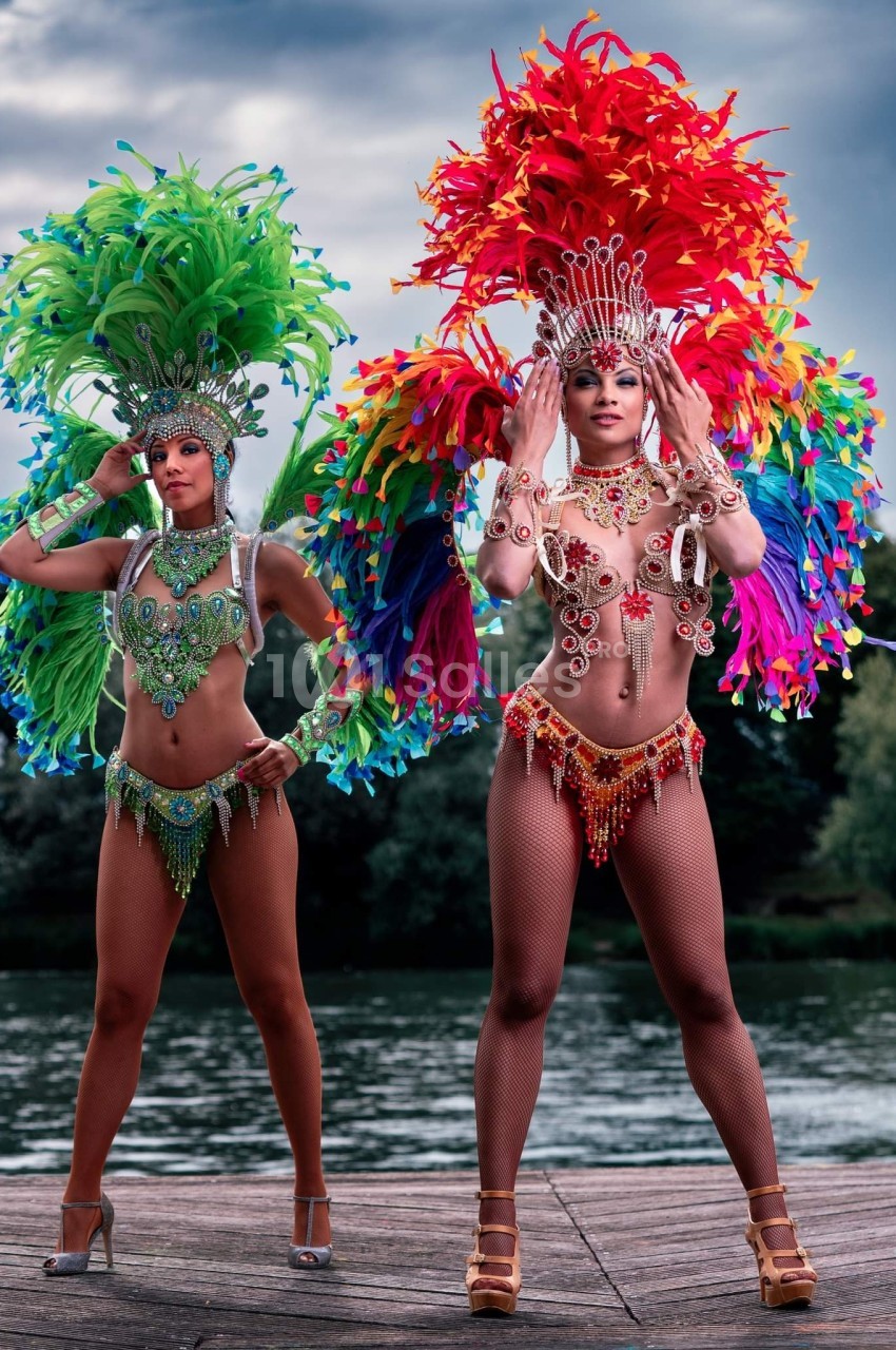 Deux danseuses en costumes colorés avec plumes, posant devant un plan d'eau et des arbres en arrière-plan.