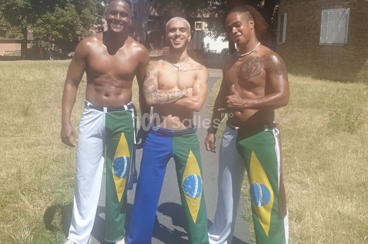 Trois hommes souriants en tenue de capoeira avec des drapeaux brésiliens, debout dans un parc ensoleillé.