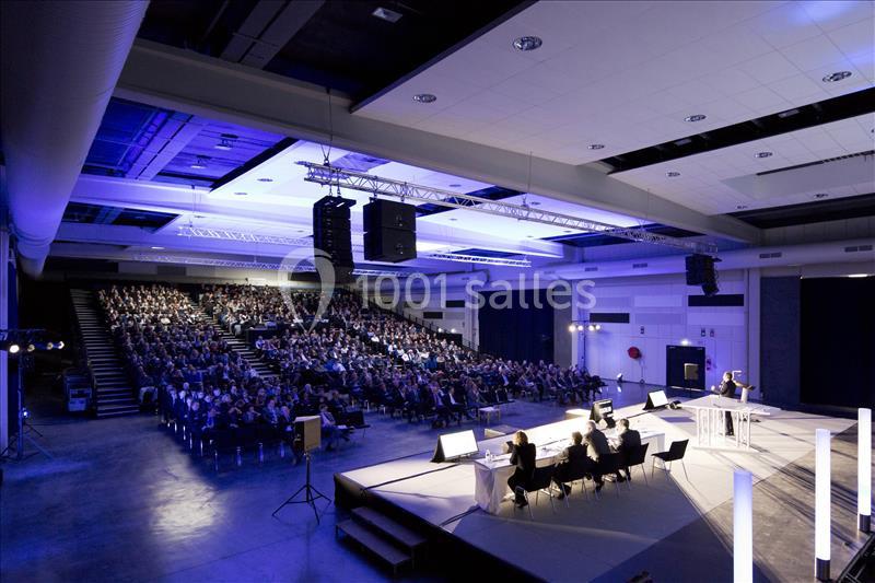 Salle de conférence moderne avec un public nombreux, une scène éclairée et un orateur s'exprimant au micro.