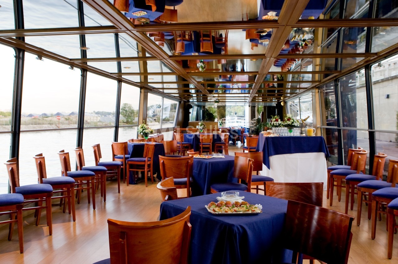 Salle de réception sur un bateau avec des tables dressées, des chaises en bois et une vue sur l'eau à travers de grandes…