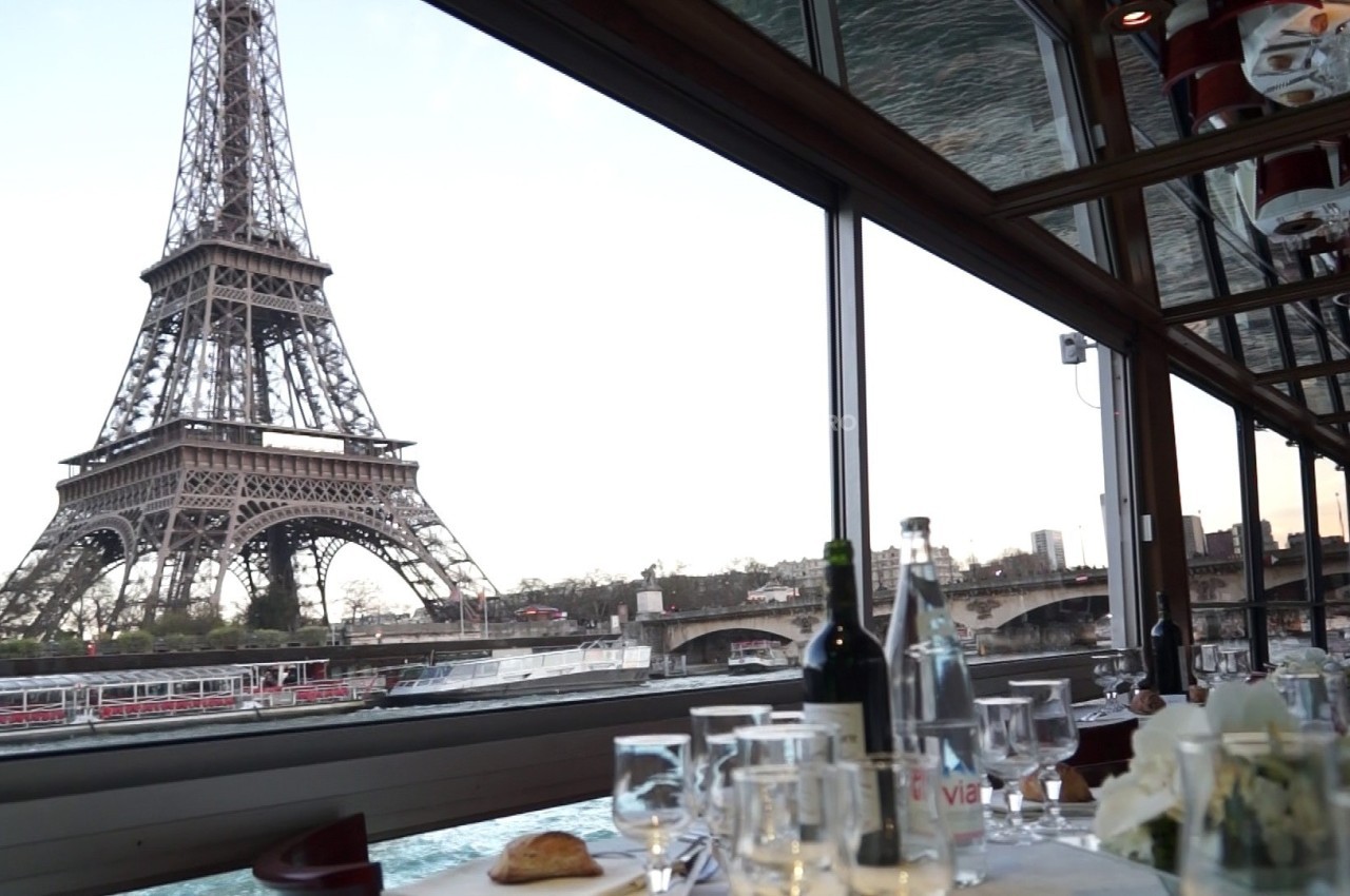 Vue de la tour Eiffel depuis l'intérieur d'un restaurant avec une table dressée et des baies vitrées.