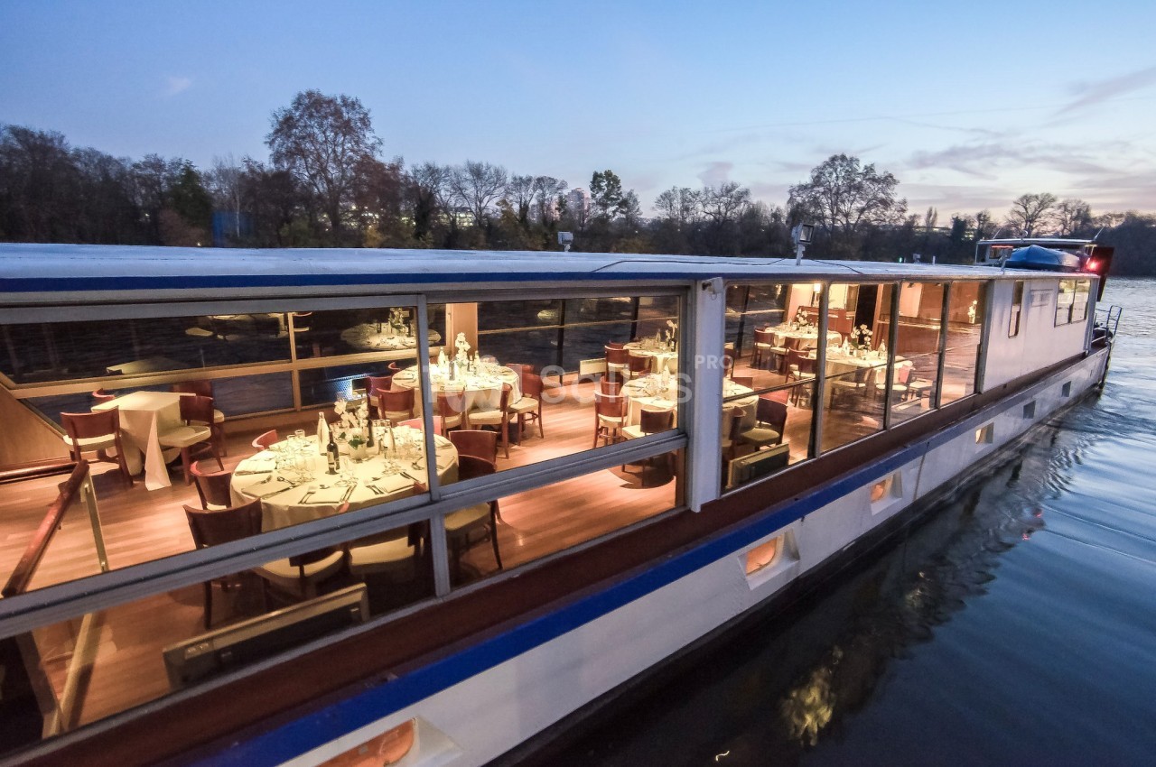 Bateau-restaurant éclairé naviguant sur une rivière au crépuscule, avec des tables dressées visibles à travers les fenêtres.