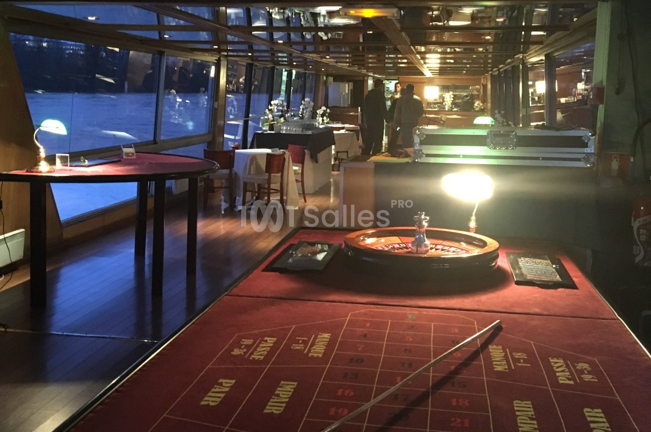 Table de roulette éclairée dans une salle de réception avec baies vitrées et tables dressées en arrière-plan.