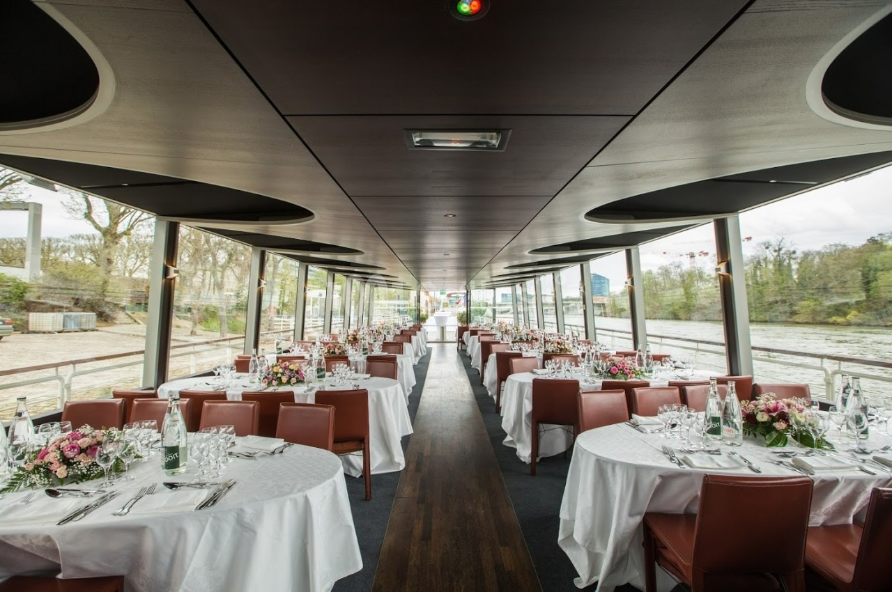 Salle de réception sur une péniche avec tables rondes dressées, nappes blanches et vue sur un paysage fluvial.