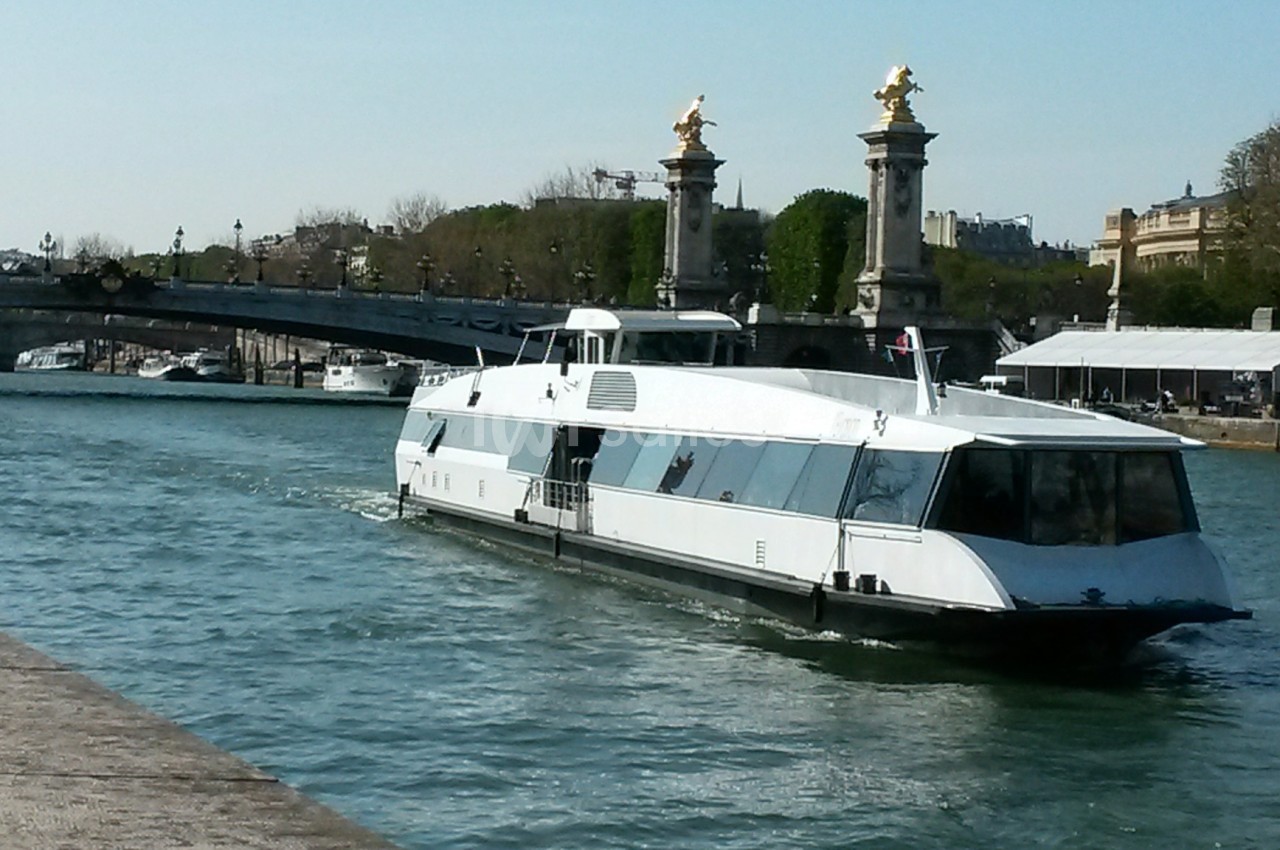 Bateau moderne naviguant sur une rivière avec un pont orné de statues dorées en arrière-plan.