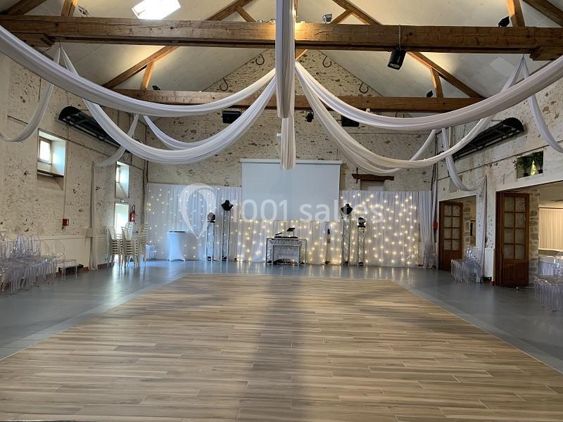 Salle de réception avec poutres apparentes, décorée de voilages blancs et éclairée par des guirlandes lumineuses.