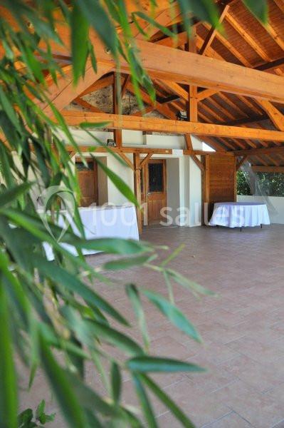 Salle avec charpente en bois apparente, tables rondes recouvertes de nappes blanches et feuillage au premier plan.