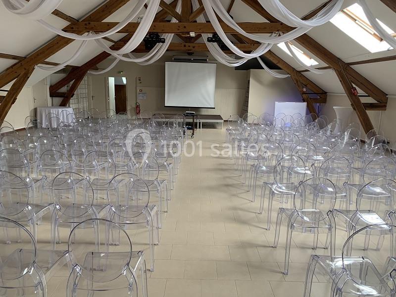 Salle de conférence avec des chaises transparentes alignées face à un écran de projection sous une charpente en bois.