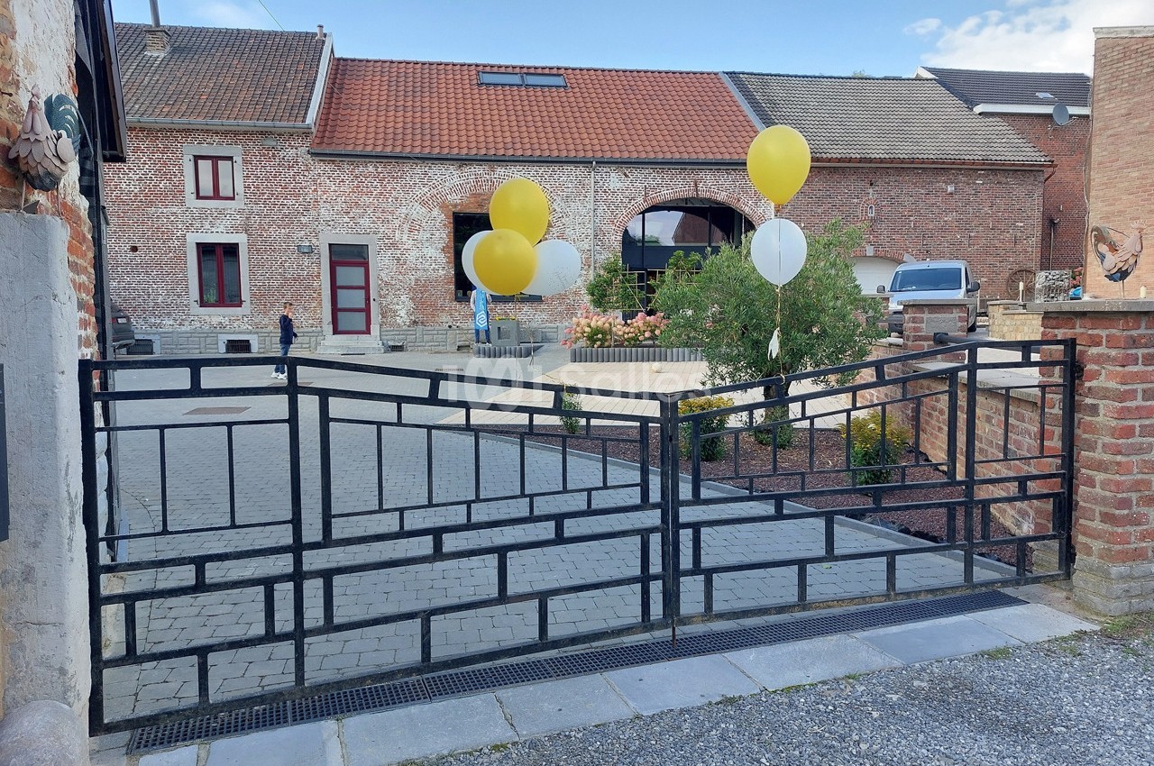 Portail métallique noir ouvert devant une cour pavée avec des ballons jaunes et blancs attachés à une clôture.