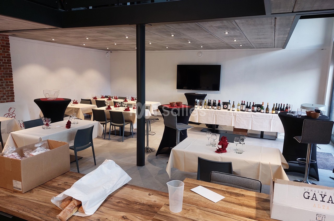 Salle aménagée pour un événement avec tables dressées, bouteilles de vin alignées et écran mural.