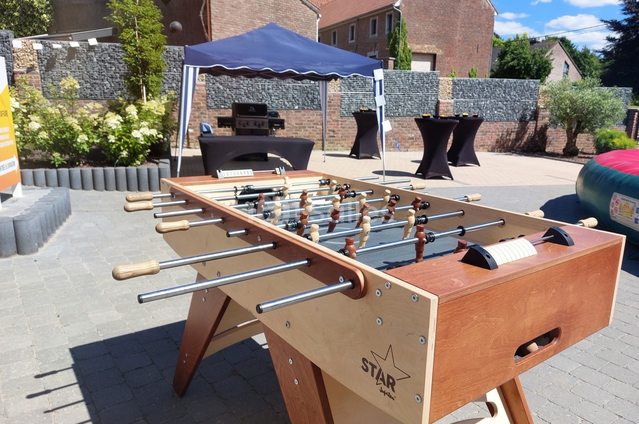 Table de baby-foot en bois installée en extérieur sur une terrasse pavée, avec un chapiteau et des tables en arrière-plan.