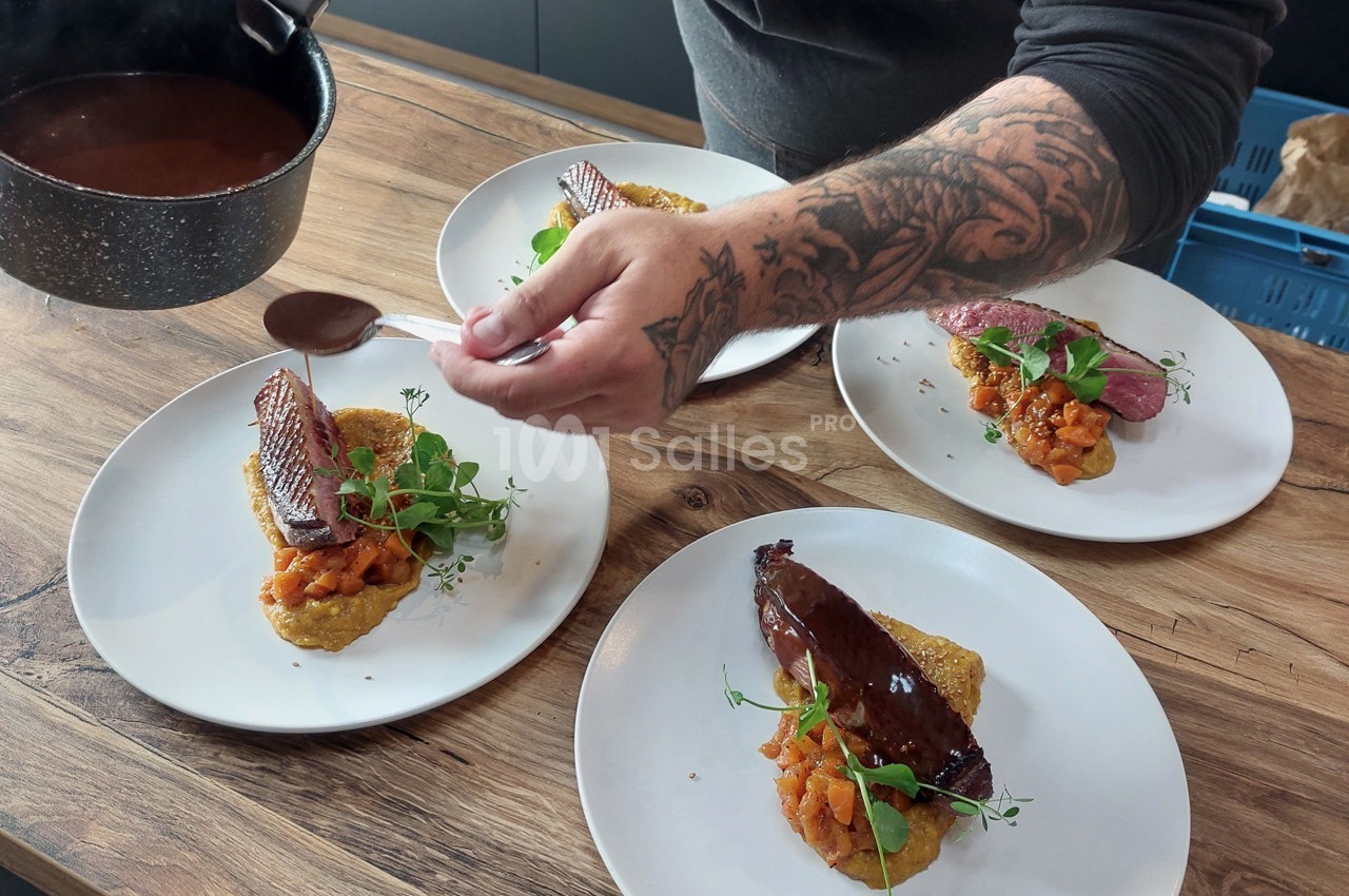 Un chef ajoute une sauce sur des assiettes garnies de viande, purée et légumes, disposées sur une table en bois.
