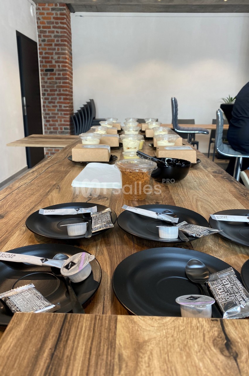 Table en bois dressée avec des assiettes noires, couverts, condiments et boissons, dans une salle aux murs en briques.