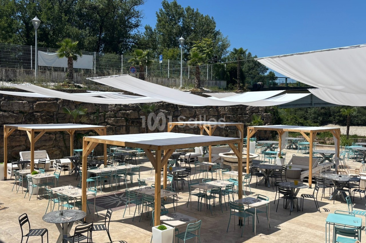 Terrasse extérieure avec tables et chaises sous des pergolas en bois et voiles d'ombrage, entourée de verdure.