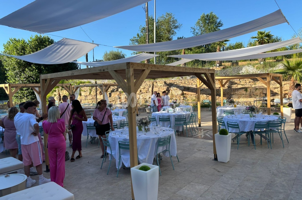 Terrasse extérieure aménagée avec des tables dressées sous des pergolas en bois, entourée de végétation et de convives.