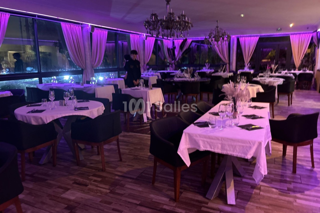 Salle de restaurant élégante avec tables dressées, nappes blanches, éclairage violet et grandes baies vitrées.