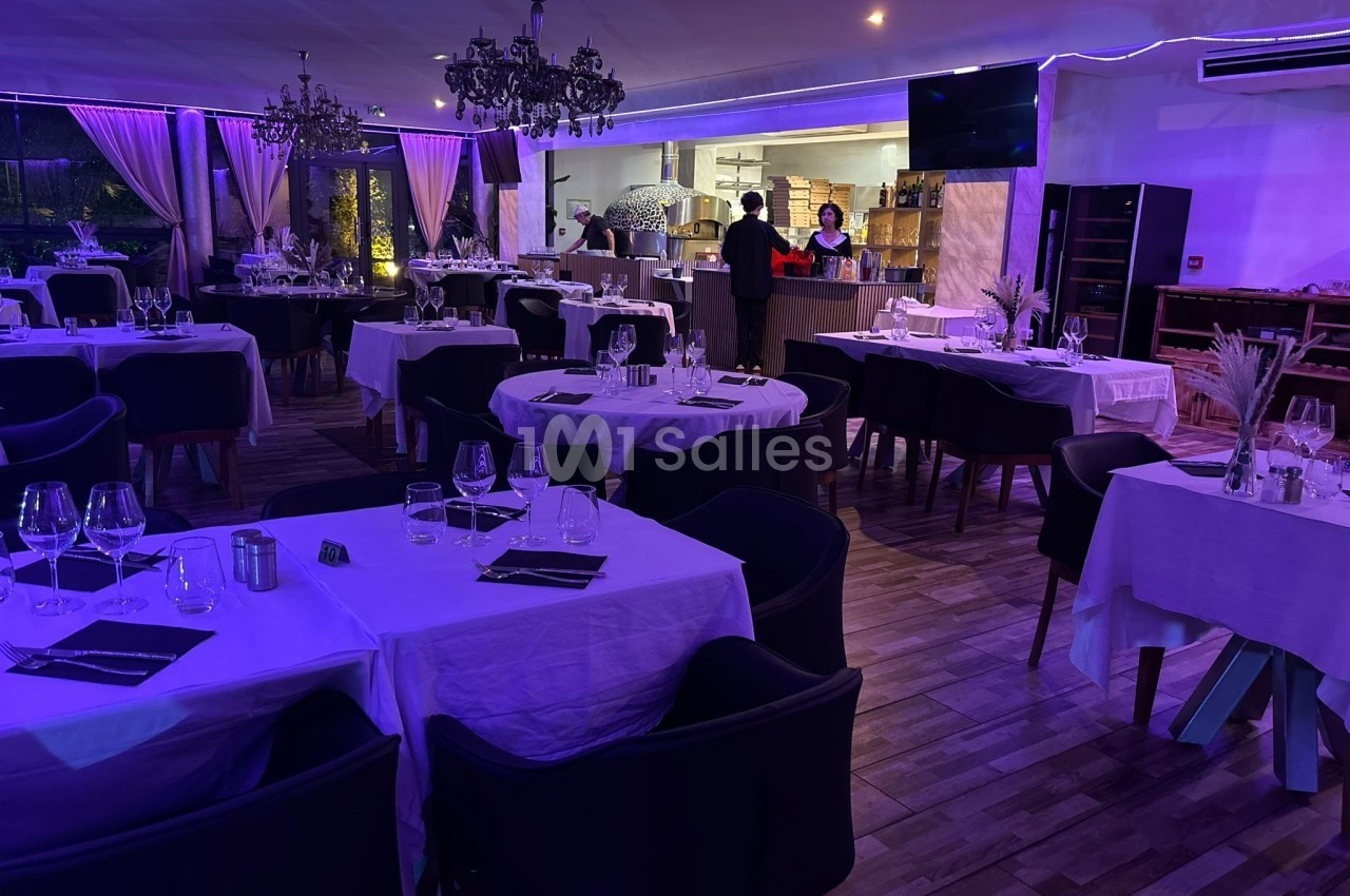 Salle de restaurant élégante avec tables dressées, éclairage tamisé violet et vue sur une cuisine ouverte.