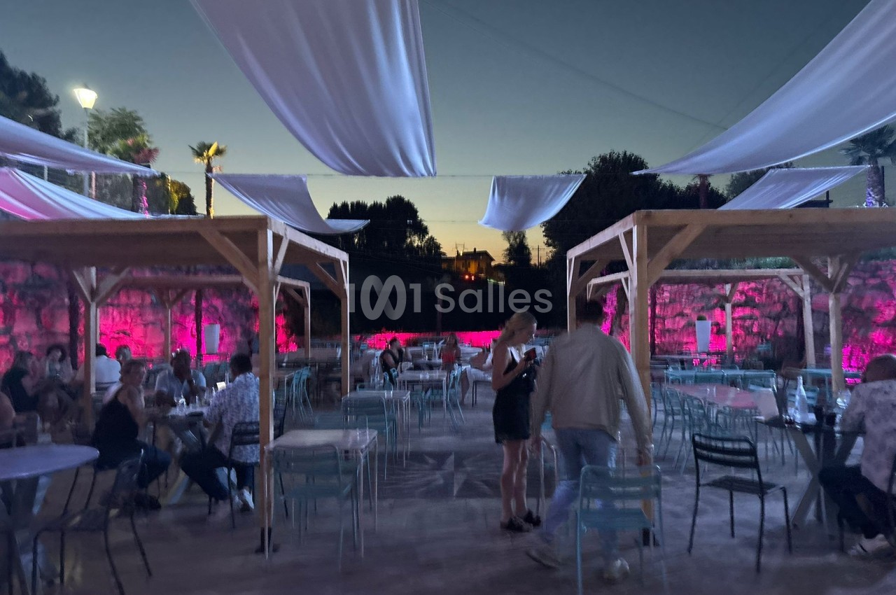 Terrasse extérieure en soirée avec des tables occupées, des voiles suspendues et un éclairage rose en arrière-plan.