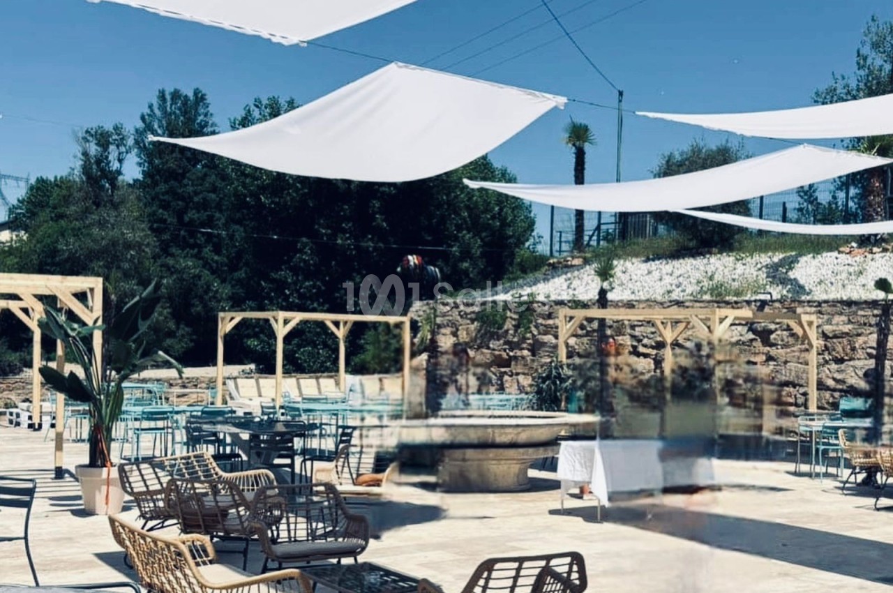 Terrasse extérieure avec des voiles d'ombrage blanches, mobilier en rotin et tables près d'une fontaine centrale.