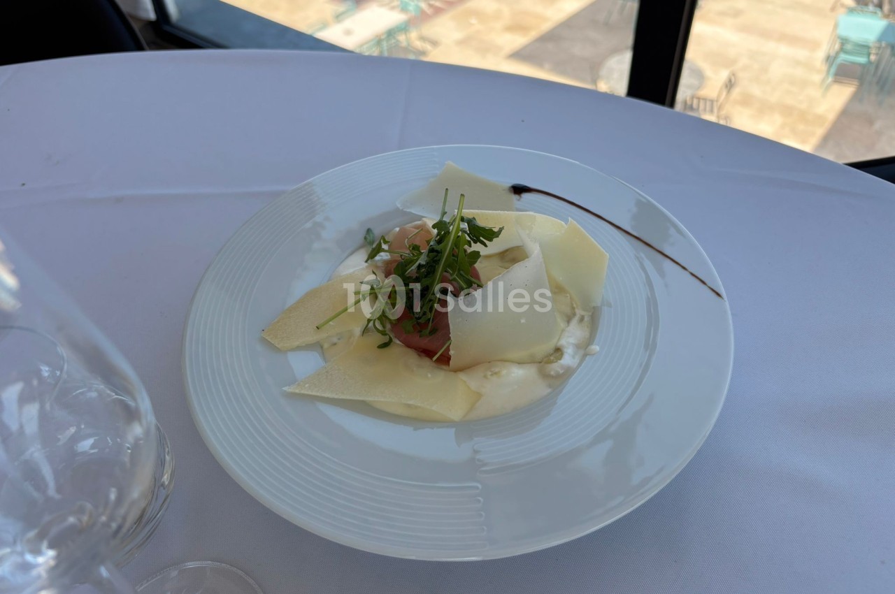 Assiette de carpaccio avec copeaux de parmesan, roquette et filet de sauce, servie sur une table blanche près d'une fenêtre.