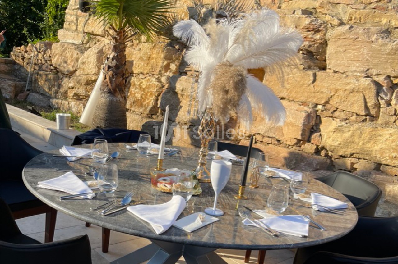 Table ronde dressée avec vaisselle élégante, bougies et décoration à plumes, en extérieur devant un mur de pierre.