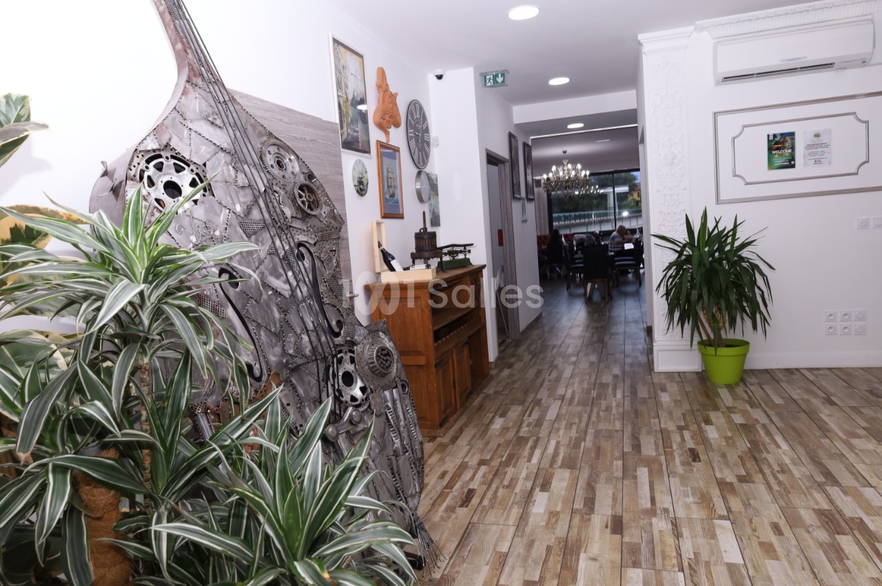 Couloir lumineux avec sol en bois, plantes vertes, sculptures métalliques et vue sur une salle à manger au fond.