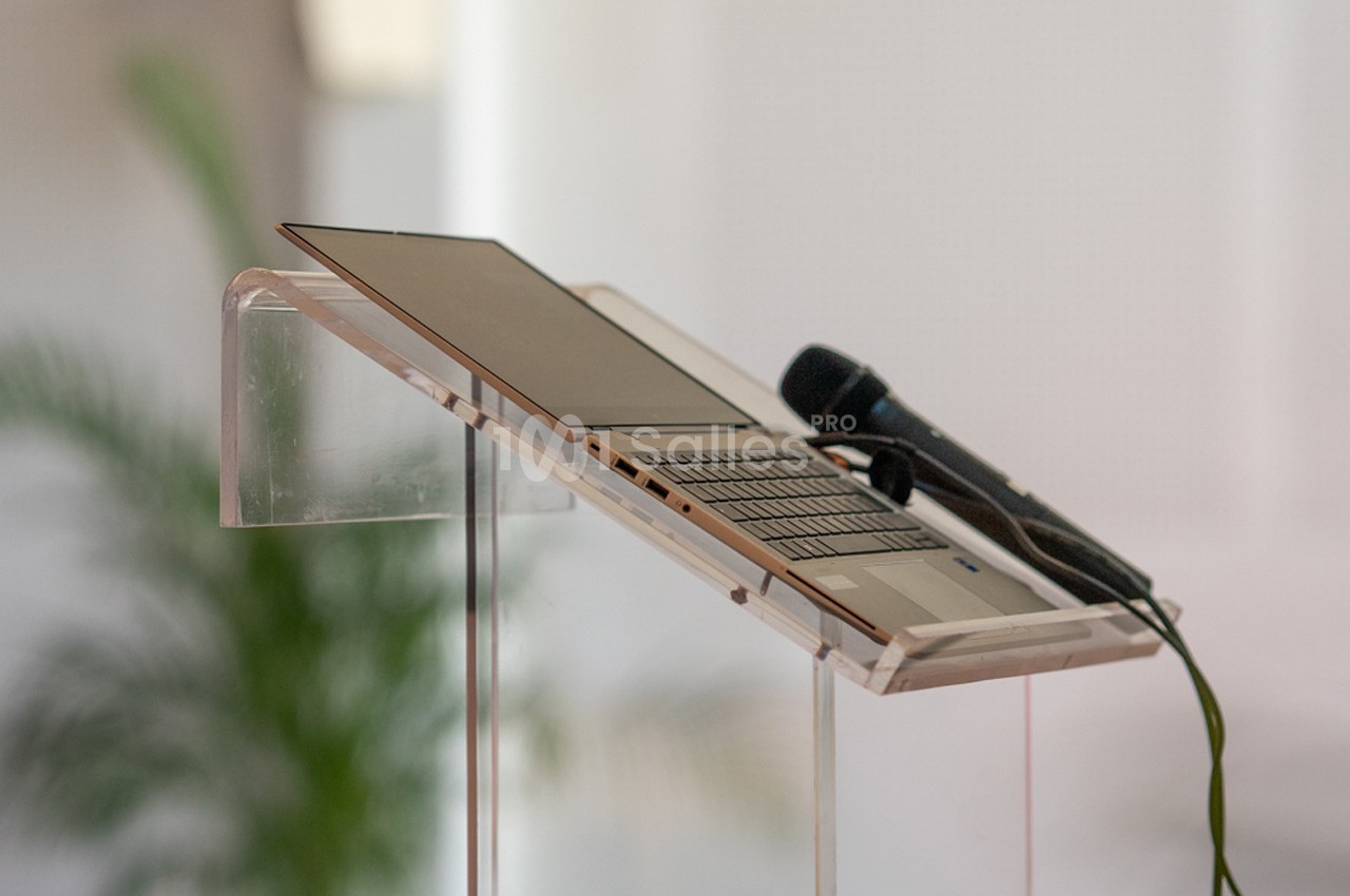 Ordinateur portable ouvert et microphone posé sur un pupitre transparent, avec une plante floue en arrière-plan.