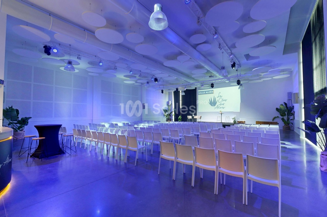 Salle de conférence moderne avec rangées de chaises blanches, éclairage bleu et écran de projection allumé.
