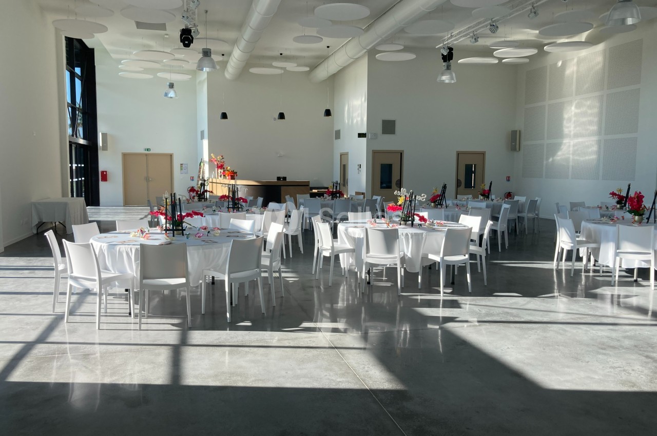 Salle lumineuse avec tables rondes dressées, chaises blanches et décorations florales colorées, prête pour un événement.